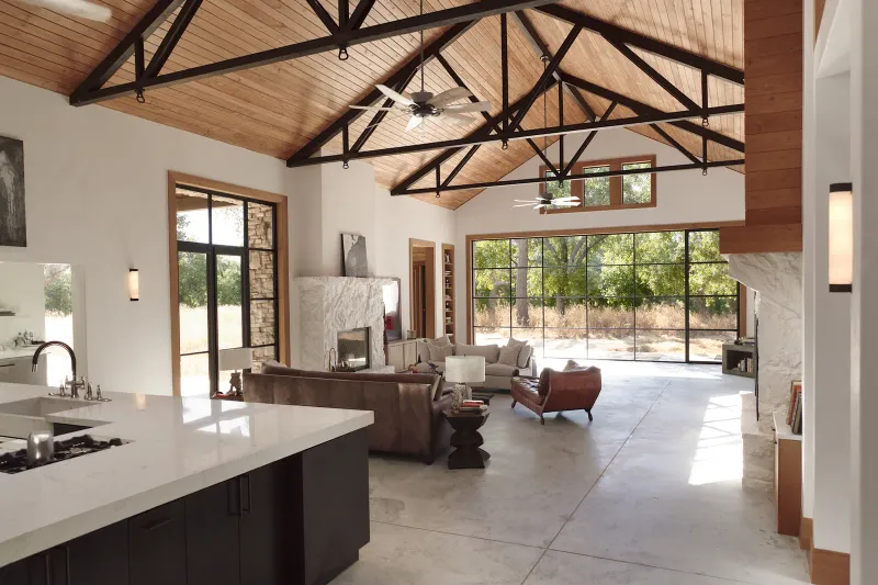 A barndominium great room with exposed black steel trusses under a wood ceiling, a wall of black steel-framed glass onto live oaks and dry grass, a limestone fireplace, polished concrete and an open kitchen island.