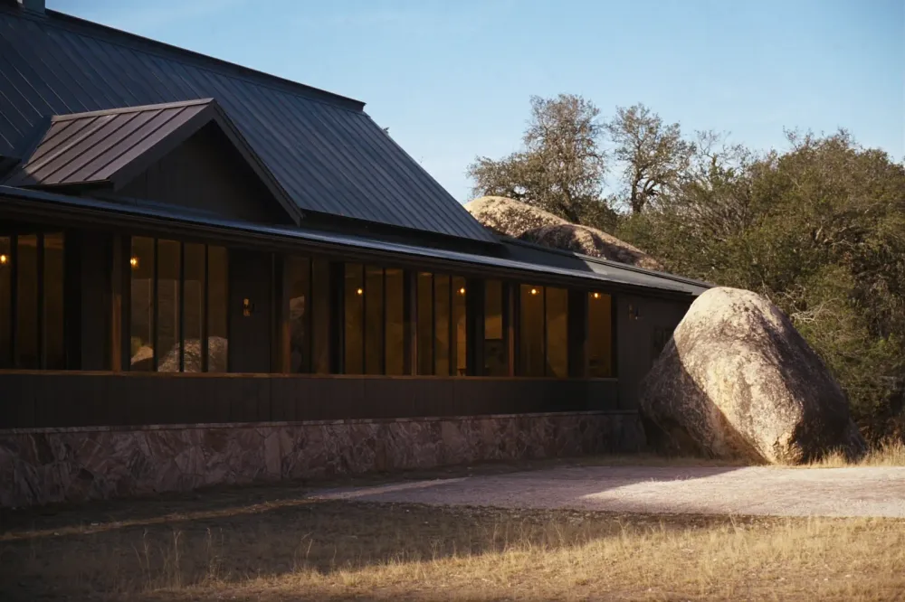 A finished barndominium with a charcoal standing seam roof, dark board and batten siding and a pink granite base, a granite boulder and live oaks behind it, Llano Uplift country.