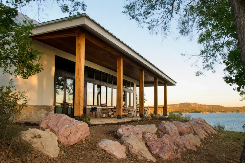 The back porch of a finished barndominium at golden hour, heavy cedar posts under a standing seam roof, black-framed glass behind, pink granite boulders in front and blue lake water below.