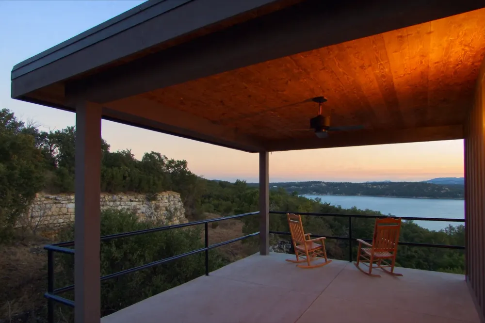 Two rocking chairs on a covered concrete porch at evening, looking over a limestone ledge and live oaks to a wide lake and low wooded hills.