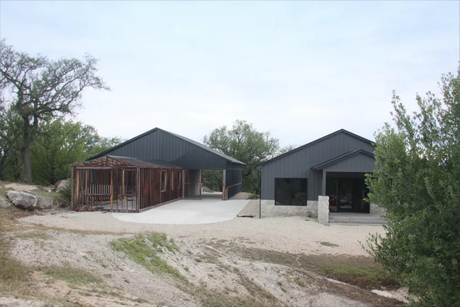A dried-in steel barndominium shell with open window bays beside its finished twin with limestone wainscot and black windows, on a limestone and cedar hillside.