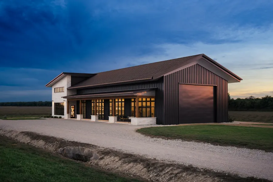 A finished barndominium at dusk with a limestone porch, tall black windows lit from inside and an attached shop bay with a roll-up door, a gravel drive crossing a culvert in front, Blackland Prairie.