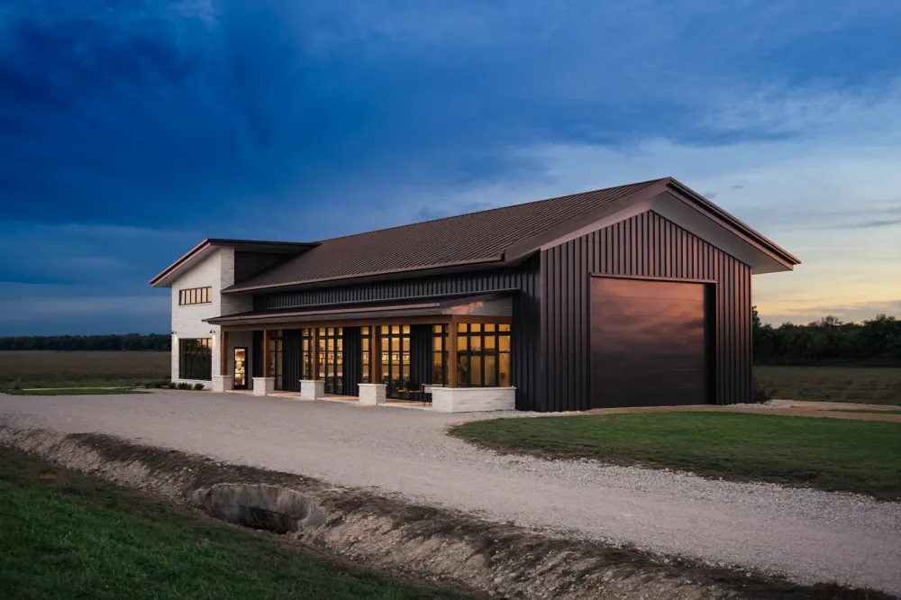 A finished barndominium at dusk with a limestone porch, tall black windows lit from inside and an attached shop bay with a roll-up door, a gravel drive crossing a culvert in front, Blackland Prairie.