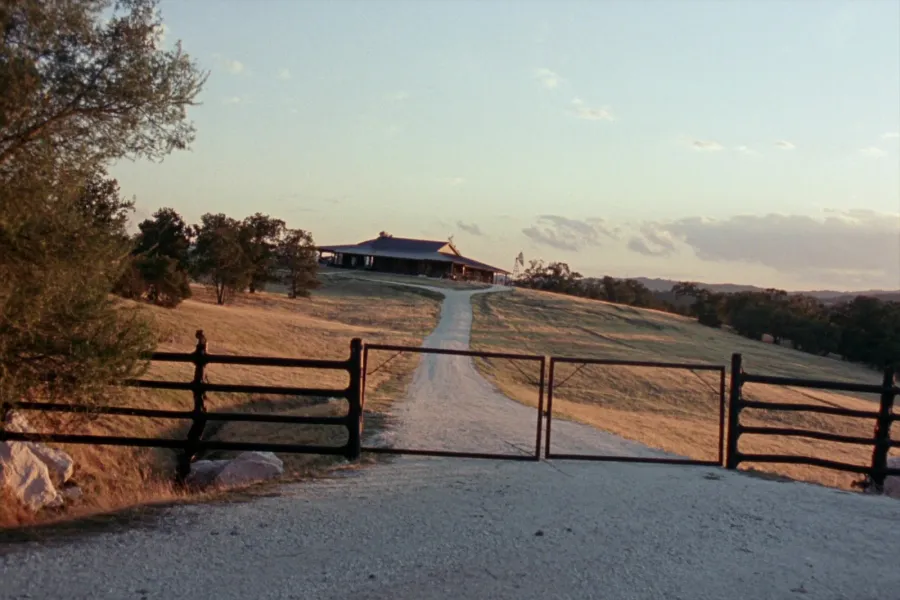 A pipe fence and double swing gate across a caliche road, with a barndominium on the rise beyond among live oaks at evening.